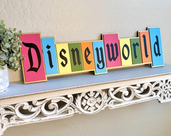 May include: A colorful wooden sign that spells out "Disneyworld" in black gothic font. The letters are on individual rectangular blocks in various colors, including pink, blue, yellow, green, and orange. The sign is displayed on a decorative shelf.