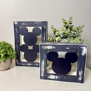 May include: Two black wooden frames with a distressed white paint finish. Each frame features a cutout of Mickey Mouse's head in black.