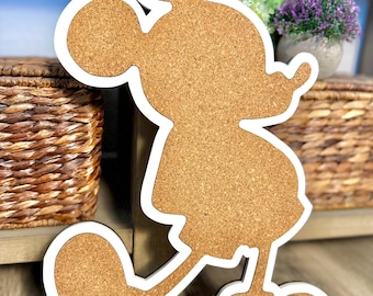 May include: A Mickey Mouse-shaped corkboard with a white border. The corkboard is in the shape of the iconic character, with a light brown cork surface. The white border outlines the shape. The corkboard is standing upright.
