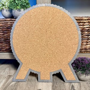 May include: A round corkboard with a gray wooden frame. The frame has a scalloped edge and a unique base design. The corkboard is displayed on a wooden surface, with decorative plants and baskets in the background.