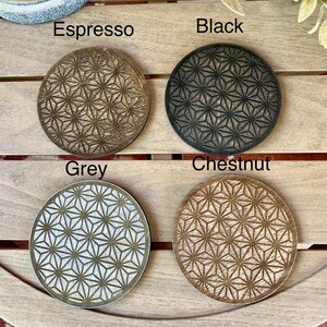May include: Four round coasters in espresso, black, grey, and chestnut. Each coaster features a geometric starburst pattern. The names of the colors are printed above each coaster. The coasters are displayed on a wooden surface.