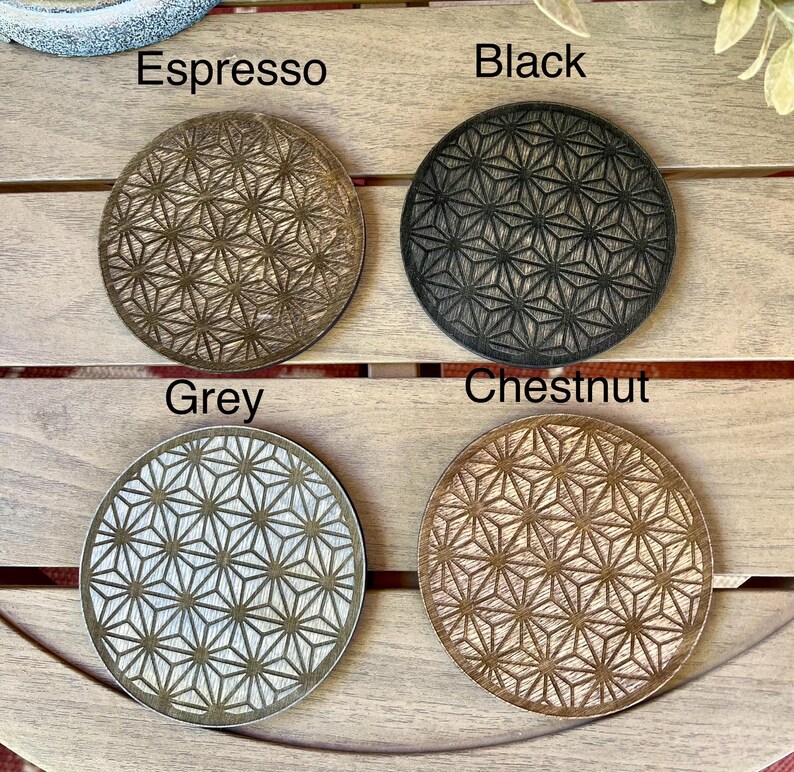 May include: Four round coasters in espresso, black, grey, and chestnut. Each coaster features a geometric starburst pattern. The names of the colors are printed above each coaster. The coasters are displayed on a wooden surface.