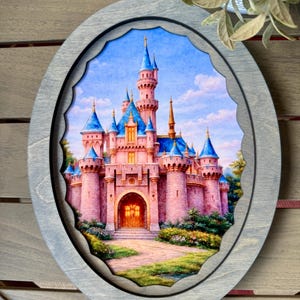 May include: An oval-shaped wooden frame with a detailed painting of a pink castle with blue turrets. The castle is set against a blue sky with clouds. The frame has a gray finish and a scalloped inner edge.