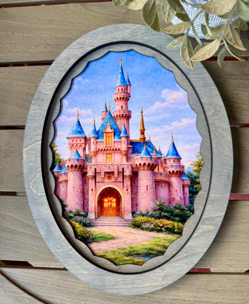 May include: An oval-shaped wooden frame with a detailed painting of a pink castle with blue turrets. The castle is set against a blue sky with clouds. The frame has a gray finish and a scalloped inner edge.