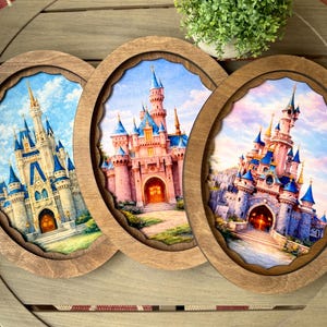May include: Three wooden framed art pieces featuring watercolor paintings of fairytale castles. Each castle has a unique design with blue and pink turrets, set against a blue sky. The frames have a scalloped edge.