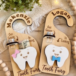 May include: Two light-colored wooden door hangers personalized with the names Makenzie and Logan. Each features a cartoon tooth, a small glass vial with a silver lid, and text reading Out Going, In Coming, and Tooth Fairy. One tooth has a pink bow.