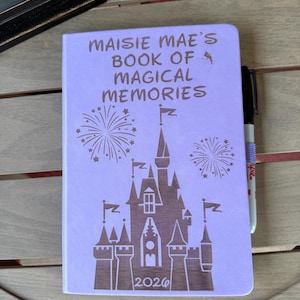 K&ouml;nnte beinhalten: Ein hellviolettes Notizbuch mit einer dunkelbraunen Gravur, die ein Schloss, Feuerwerk und den Text "MAISIE MAE'S BOOK OF MAGICAL MEMORIES" &uuml;ber dem Jahr "2026" zeigt. Ein schwarzer Stift steckt an der Seite des Notizbuchumschlags.