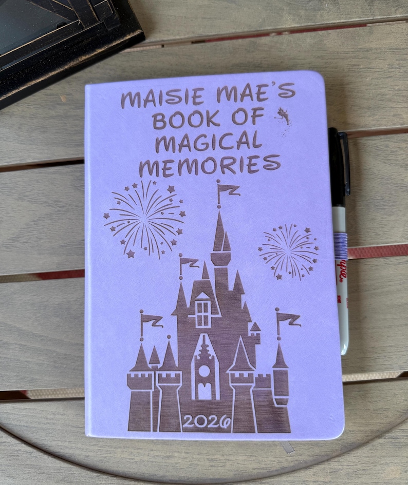 K&ouml;nnte beinhalten: Ein hellviolettes Notizbuch mit einer dunkelbraunen Gravur, die ein Schloss, Feuerwerk und den Text "MAISIE MAE'S BOOK OF MAGICAL MEMORIES" &uuml;ber dem Jahr "2026" zeigt. Ein schwarzer Stift steckt an der Seite des Notizbuchumschlags.