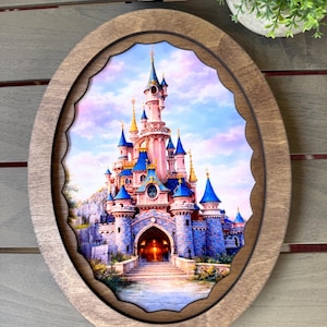 May include: An oval wooden frame surrounds a colorful image of a fairytale castle with blue turrets and a pink facade. The castle is set against a pastel sky, with a scalloped edge around the image.