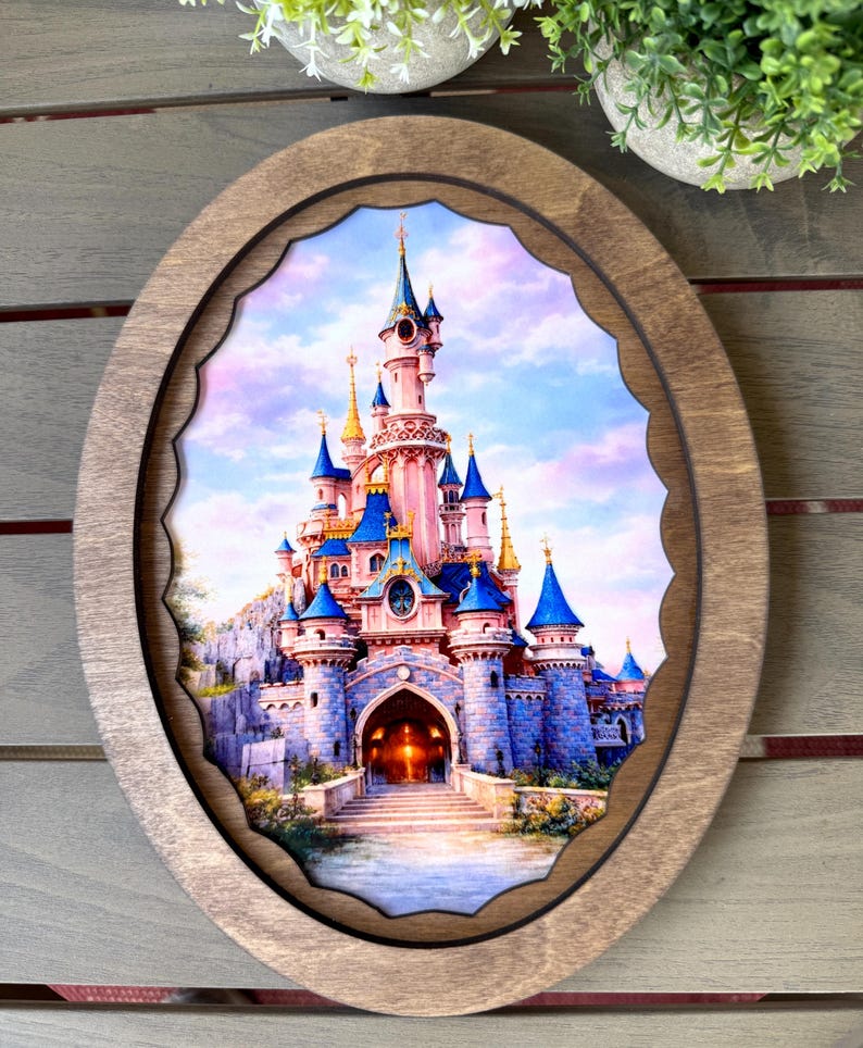 May include: An oval wooden frame surrounds a colorful image of a fairytale castle with blue turrets and a pink facade. The castle is set against a pastel sky, with a scalloped edge around the image.