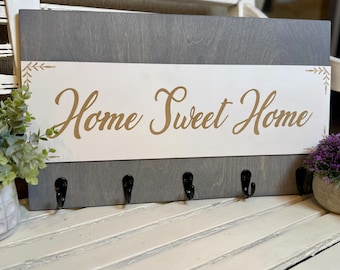 May include: A gray wooden key holder with the phrase "Home Sweet Home" in gold script. The key holder has five black hooks and decorative leaf accents. The key holder is a decorative home decor item.