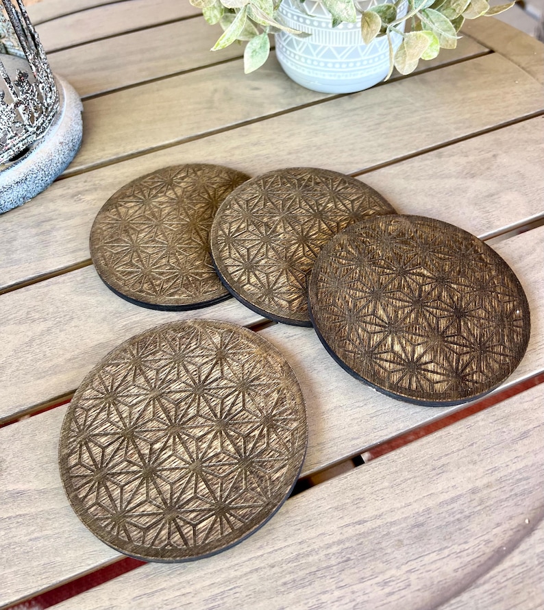May include: Four round, wooden coasters with a geometric pattern. The coasters are a warm brown color with a dark brown border. The coasters are arranged on a light-colored wooden surface.