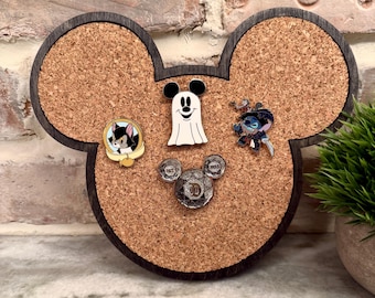 May include: A Mickey Mouse-shaped corkboard with a dark wood border. The corkboard displays several enamel pins, including a black and white cat, a ghost, and a blue alien. A small green plant is visible in the background.