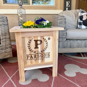 May include: A wooden planter box with the text "the PARSONS family" and the letter "P" surrounded by a laurel wreath. The planter is on a red and white rug with a Mickey Mouse design. The planter is filled with yellow, purple, and white flowers.