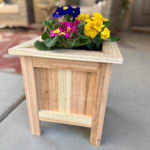 May include: A wooden planter box filled with colorful flowers. The flowers are a mix of yellow, pink, and blue. The planter is made of light-colored wood and has a rectangular shape. The planter is sitting on a concrete surface.
