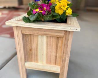 May include: A wooden planter box filled with colorful flowers. The flowers are a mix of yellow, pink, and blue. The planter is made of light-colored wood and has a rectangular shape. The planter is sitting on a concrete surface.