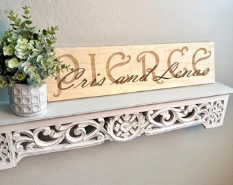 May include: A wooden sign with the text "Pierce Chris and Lenae" engraved in a cursive font. The sign is light brown and has a natural wood grain. It is sitting on a white shelf with intricate carvings.