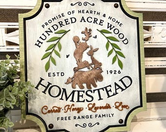 May include: Wooden sign with the text "Hundred Acre Wood Homestead" and the words "Promise of Hearth & Home" and "Free Range Family". The sign features a light green border, a distressed white background, and wooden cutouts of animals and leaves.