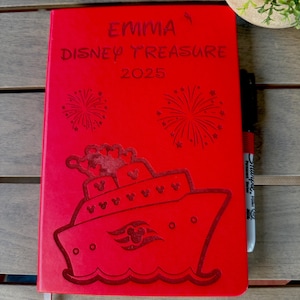 K&ouml;nnte beinhalten: Ein leuchtend rotes, rechteckiges Notizbuch mit einer Gravur eines Kreuzfahrtschiffes mit Mickey-Mouse-Symbol auf dem Rumpf, zwei Feuerwerken und dem Text EMMA DISNEY TREASURE 2025. Ein schwarzer Permanentmarker liegt an der rechten Kante.