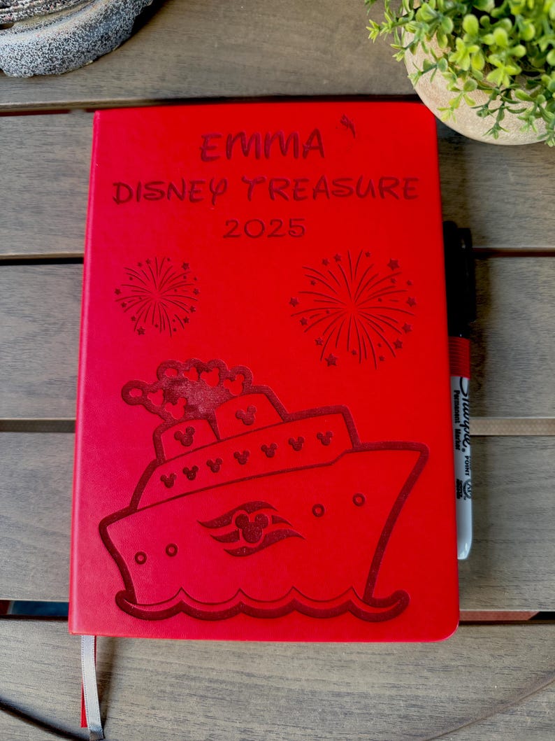 K&ouml;nnte beinhalten: Ein leuchtend rotes, rechteckiges Notizbuch mit einer Gravur eines Kreuzfahrtschiffes mit Mickey-Mouse-Symbol auf dem Rumpf, zwei Feuerwerken und dem Text EMMA DISNEY TREASURE 2025. Ein schwarzer Permanentmarker liegt an der rechten Kante.