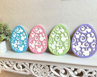 May include: Four wooden Easter eggs decorated with a white swirl pattern and Mickey Mouse silhouettes. The eggs are painted in light blue, pink, green, and purple.