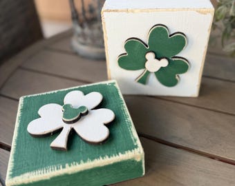Könnte beinhalten: Zwei Holzblöcke mit grünen Kleeblatt-Designs. Ein Block ist weiß mit einem grünen Kleeblatt und einem weißen Mickey-Maus-Kopf. Der andere Block ist grün mit einem weißen Kleeblatt und einem grünen Mickey-Maus-Kopf.