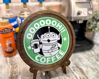 May include: A wooden round sign with a green background and a white alien character holding a cup of coffee. The sign says "Oooooh! Coffee" in black text.