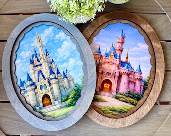 May include: Two oval wooden framed prints featuring fairytale castles. One frame is gray, the other is brown. The castle art has blue, pink, and gold accents. The background shows a blue sky with clouds and green trees.