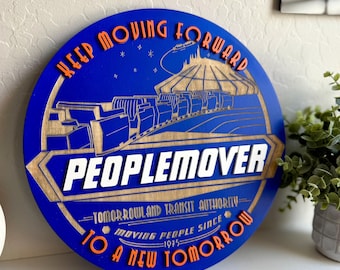 May include: A blue circular wooden sign with the text "Keep Moving Forward" in orange letters. The sign features a stylized image of the Peoplemover ride at Disney World's Tomorrowland, with the text "Peoplemover" in white letters. The sign also includes the text "Tomorrowland Transit Authority" and "Moving People Since 1975" in smaller white letters.