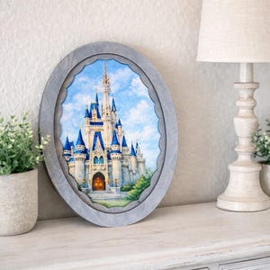 May include: An oval-shaped framed print of a fairytale castle with blue turrets and a gold-colored door. The frame is gray and has a scalloped edge. The artwork is displayed on a white surface with a lamp and greenery in the background.