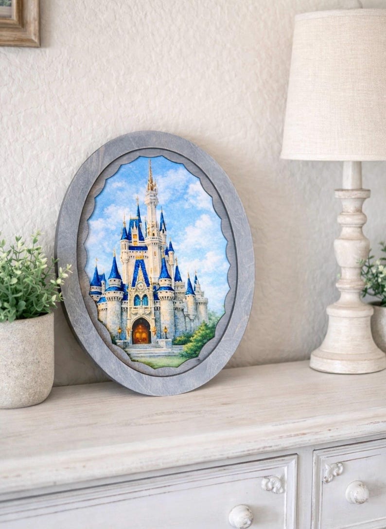 May include: An oval-shaped framed print of a fairytale castle with blue turrets and a gold-colored door. The frame is gray and has a scalloped edge. The artwork is displayed on a white surface with a lamp and greenery in the background.