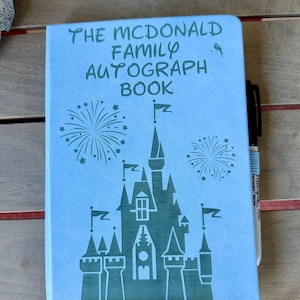 K&ouml;nnte beinhalten: Ein hellblaues Autogrammbuch mit einer dunkelgr&uuml;nen Grafik eines Schlosses und Feuerwerksk&ouml;rpern. Der Text auf dem Einband lautet THE MCDONALD FAMILY AUTOGRAPH BOOK. Ein schwarzer Permanentmarker liegt an der rechten Seite des rechteckigen Buches.