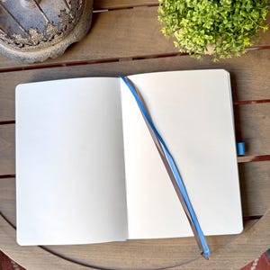 K&ouml;nnte beinhalten: Ein aufgeschlagenes Notizbuch mit leeren wei&szlig;en Seiten und einem hellblauen Lesezeichenband. Das Notizbuch verf&uuml;gt &uuml;ber eine stabile Bindung und eine passende blaue elastische Stiftschlaufe an der Seite f&uuml;r eine saubere Schreibfl&auml;che.