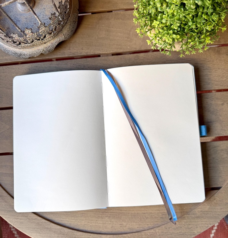 K&ouml;nnte beinhalten: Ein aufgeschlagenes Notizbuch mit leeren wei&szlig;en Seiten und einem hellblauen Lesezeichenband. Das Notizbuch verf&uuml;gt &uuml;ber eine stabile Bindung und eine passende blaue elastische Stiftschlaufe an der Seite f&uuml;r eine saubere Schreibfl&auml;che.