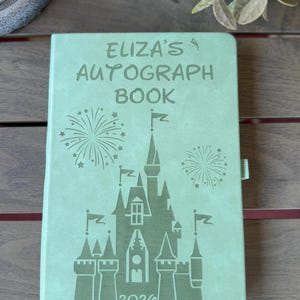 K&ouml;nnte beinhalten: Ein hellgr&uuml;nes Autogrammbuch mit einer Gravur eines Schlosses und Feuerwerk sowie dem Text "ELIZA'S AUTOGRAPH BOOK" und "2026" auf dem Einband. Das Notizbuch hat eine glatte, matte Oberfl&auml;che und eine kleine Lesezeichenlasche aus Stoff.