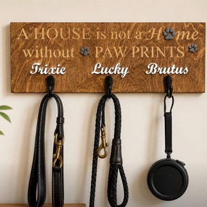 May include: A rectangular wooden wall-mounted leash holder with a dark brown stained finish. The text reads A HOUSE is not a Home without PAW PRINTS, with the names Trixie, Lucky, and Brutus below. Three black metal hooks hold pet leashes.