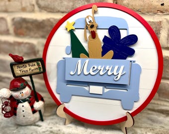 May include: A round, wooden Christmas decoration with a red border and a white background. The design features a light blue truck carrying a cartoon dog, a Christmas tree, and a wrapped gift. The word "Merry" is displayed in white. A snowman figurine is on the left.