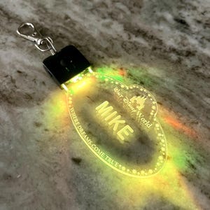 May include: A clear plastic light-up name tag with the text "Walt Disney World" and "Where dreams come true" around the edge. The name "Mike" is in the center of the tag. The tag is lit up with a yellow and green glow.