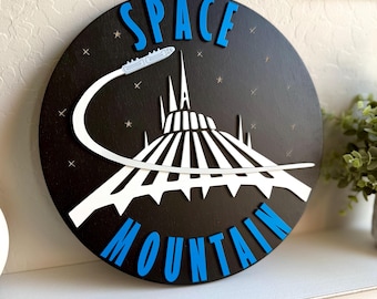 May include: A black wooden round sign with the words "SPACE MOUNTAIN" in blue letters. The sign features a white silhouette of the Space Mountain ride with a blue spaceship.