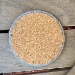 May include: Round cork coaster with a gray wooden border. The coaster has a natural cork center and a light gray wooden edge. The coaster is designed to protect surfaces from heat and moisture.