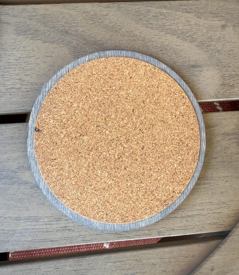 May include: Round cork coaster with a gray wooden border. The coaster has a natural cork center and a light gray wooden edge. The coaster is designed to protect surfaces from heat and moisture.