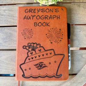 K&ouml;nnte beinhalten: Ein beigefarbenes Autogrammbuch aus leder&auml;hnlichem Material mit der Gravur GREYSON'S AUTOGRAPH BOOK, zwei Feuerwerksmotiven und einer Illustration eines Kreuzfahrtschiffes mit Mickey-Maus-Symbolen. Ein schwarzer Sharpie-Stift steckt an der rechten Seite.