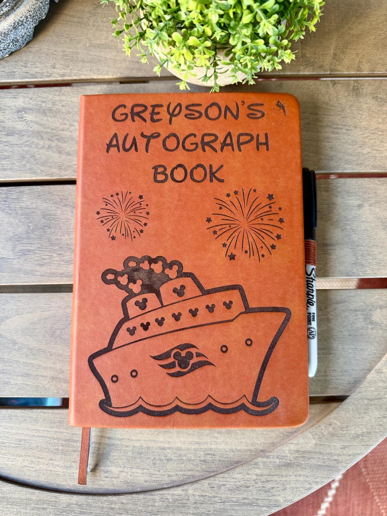 K&ouml;nnte beinhalten: Ein beigefarbenes Autogrammbuch aus leder&auml;hnlichem Material mit der Gravur GREYSON'S AUTOGRAPH BOOK, zwei Feuerwerksmotiven und einer Illustration eines Kreuzfahrtschiffes mit Mickey-Maus-Symbolen. Ein schwarzer Sharpie-Stift steckt an der rechten Seite.