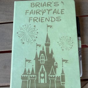 K&ouml;nnte beinhalten: Ein hellgr&uuml;nes Notizbuch mit einer Gravur eines Schlosses und Feuerwerk sowie dem Text "BRIAR'S FAIRYTALE FRIENDS" auf dem Einband. Ein schwarzer Stift steckt in einer passenden hellgr&uuml;nen elastischen Stiftschlaufe an der Seite des Notizbuchs.