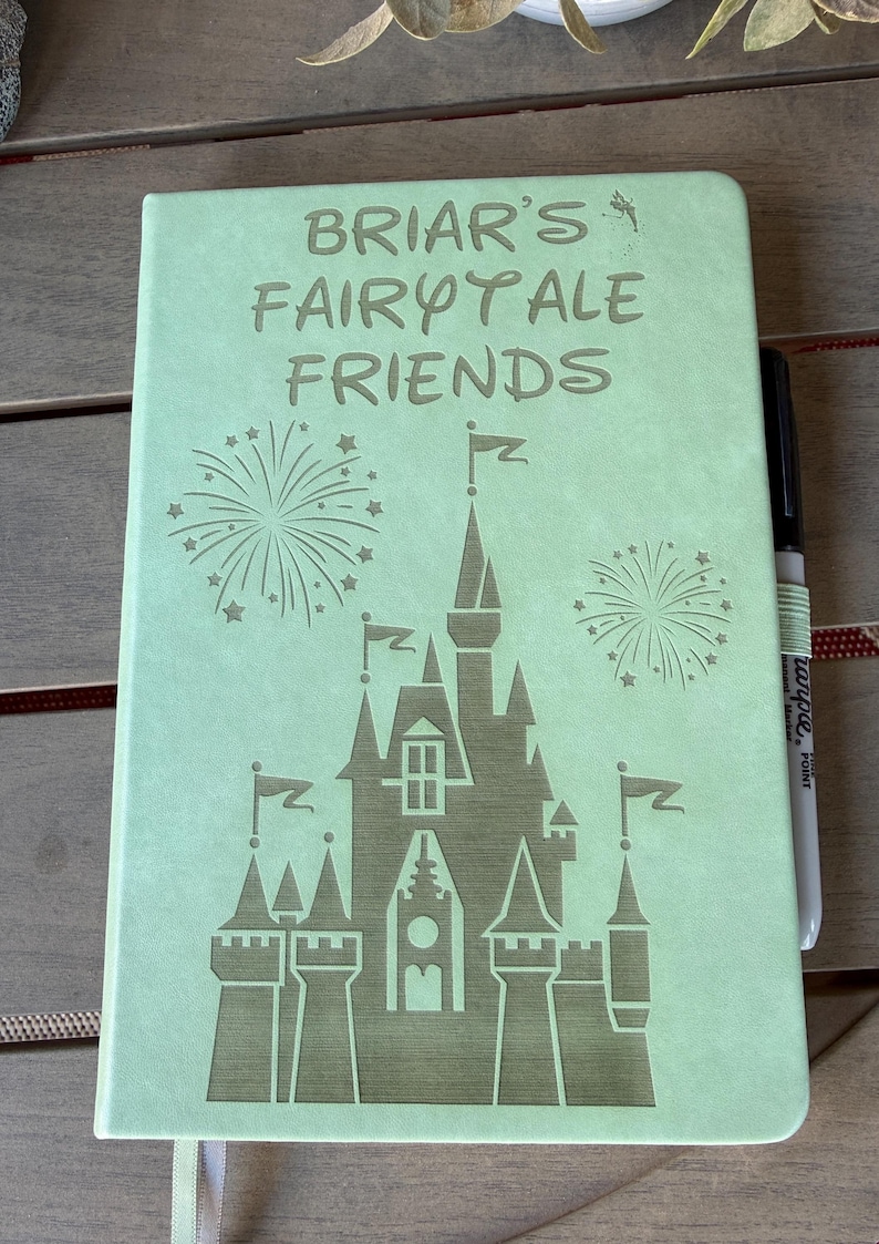 K&ouml;nnte beinhalten: Ein hellgr&uuml;nes Notizbuch mit einer Gravur eines Schlosses und Feuerwerk sowie dem Text "BRIAR'S FAIRYTALE FRIENDS" auf dem Einband. Ein schwarzer Stift steckt in einer passenden hellgr&uuml;nen elastischen Stiftschlaufe an der Seite des Notizbuchs.