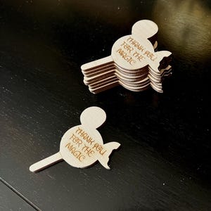 May include: A stack of light-colored wooden cutouts in the shape of a mouse head with a tail. The text "THANK YOU FOR THE MAGIC" is engraved on the head. One cutout is separate from the stack. The cutouts are on a dark surface.