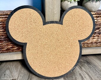 May include: A Mickey Mouse-shaped corkboard with a black wooden frame. The corkboard is light brown and the frame is dark. The board is sitting on a wooden surface.