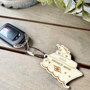 May include: A wooden keychain shaped like the Disney Wish cruise ship. The keychain is light brown and has the words "Disney Wish" and "2025" engraved on it. There are Mickey Mouse ears on the top of the ship.