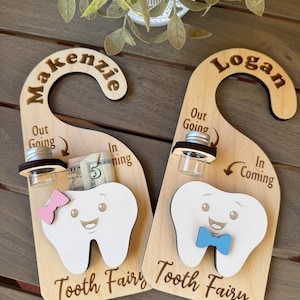 May include: Two wooden Tooth Fairy door hangers. Each has a tooth-shaped design with a smiling face, a bow tie or bow, and a small jar. One says "Makenzie" and the other says "Logan".