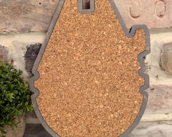 May include: A corkboard in the shape of the Millennium Falcon, with a gray wooden frame. The corkboard is a light brown color and is set against a brick wall. A small potted plant is visible on the left.